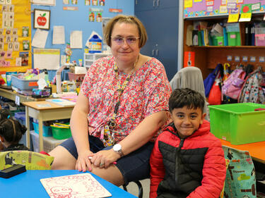 Woman with short hair, wearing glasses and floral shirt sits next to young kindergarten boy in a classroom